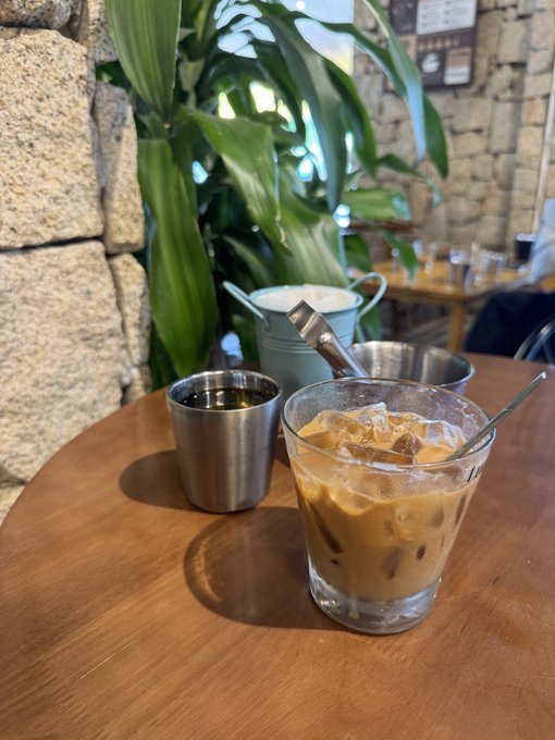 An iced coffee in a glass with a metal straw sits on a wooden table alongside a metal cup, with a green plant and stone wall in the background.