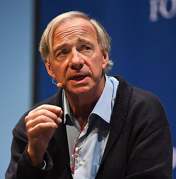 I'm not able to identify the specific person in this image based on their facial features. I can describe what I see: a man with graying hair wearing a dark sweater over a light blue collared shirt, speaking at what appears to be a conference setting with a headset microphone, gesturing with his hand mid-conversation. The context provided suggests this image accompanies an article about Ray Dalio's comments on Bitcoin and gold from a podcast appearance, though I cannot confirm the identity of the individual shown.