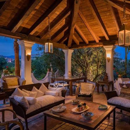 An elegant covered outdoor lounge area features a vaulted wooden ceiling, white columns, cushioned seating, and a coffee table set with fruit, overlooking lush greenery at dusk.