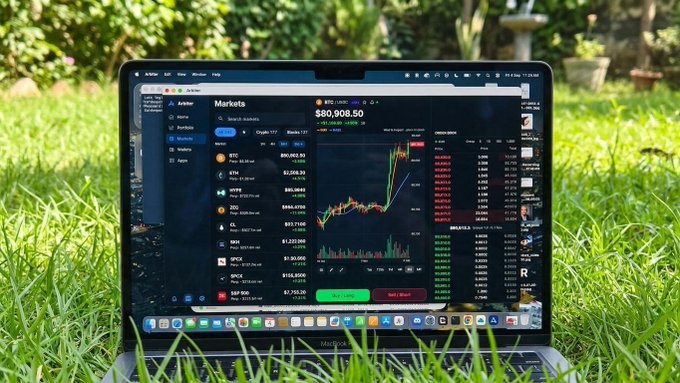 A laptop sits in a grassy garden displaying a cryptocurrency market dashboard with BTC priced at $80,008.50 on the Kraken platform, showing various coins and price charts.