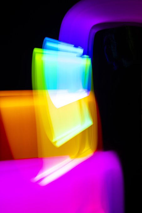 An abstract light painting or long-exposure photograph featuring overlapping colorful light shapes in yellow, orange, blue, and magenta against a dark background.