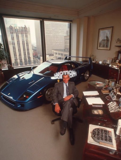 Vintage photograph of a man in a business suit sitting at a desk in a high-rise office, with a blue Ferrari F40 supercar parked behind him on the floor-to-ceiling window side overlooking a city skyline.