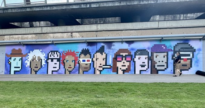 The image features a large mural painted on a wall, depicting a group of people wearing sunglasses and hats. The mural includes various characters such as a man with a hat, a woman with sunglasses, and others. A person can be seen walking past the mural in the foreground. The background of the image is a grassy area, providing a contrast to the colorful mural on the wall. The post accompanying this image mentions that the mural is part of an open-air urban art museum located in Barcelona, Spain.