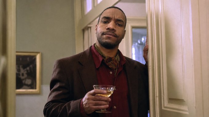 A well-dressed man holding a glass of an amber drink stands in a doorway, looking directly at the camera with a calm, composed expression. The image's intimate, contemplative mood contrasts with the post's message about refusing charity to those who don't ask, suggesting a quiet refusal or judgment toward a begging hand.