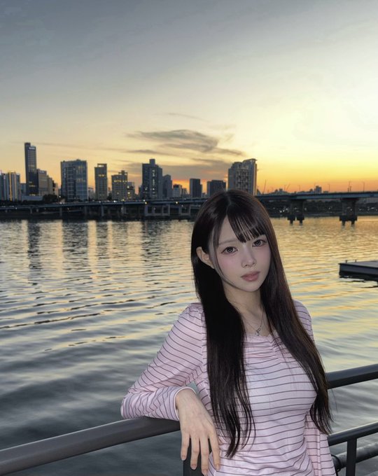 A young woman with long dark hair wearing a pink and white striped long-sleeve shirt poses at a railing by a river, with a city skyline and sunset visible in the background.