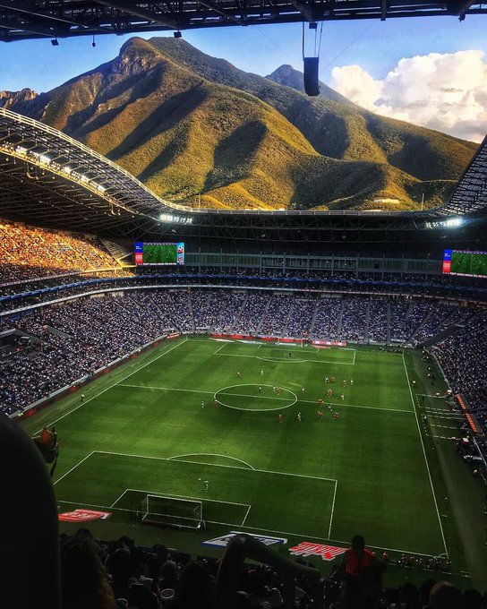 The image shows a large soccer stadium filled with spectators, with mountains visible in the background. A group of people can be seen sitting in the stands, watching the game unfold. The photo captures the excitement and atmosphere of the event as fans gather together to enjoy the match.