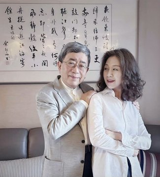 An older man with gray hair and glasses embraces a younger woman with curly dark hair as they pose together in front of a wall adorned with traditional Chinese calligraphy. The caption's mocking reference to the man being "too old" appears to critique the visible age gap between the couple in the photo.