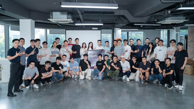 A large group of young people pose together for a group photo in a modern office space, holding a sign that reads 'Meetup'.