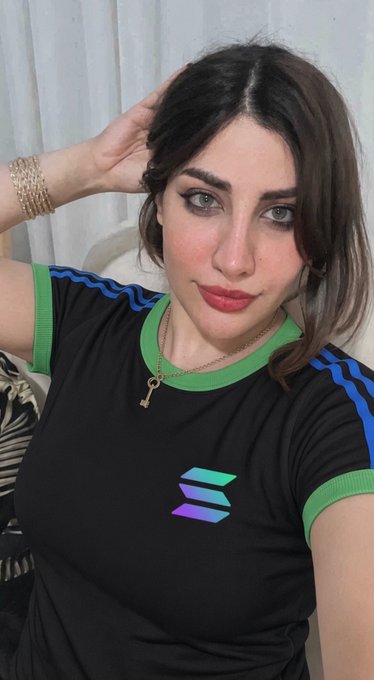 A woman with long dark hair, bold eye makeup, and red lipstick poses indoors wearing a black t-shirt with green and blue accents and a key pendant necklace.