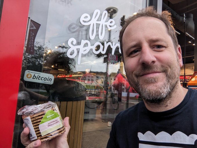 A smiling bearded man holds up a stroopwafel from Albert Heijin front of a SPAR store window advertising "Wij accepteren bitcoin" (We accept bitcoin), illustrating his purchase made via a Lightning QR code iPad payment at the Arnhem SPAR.