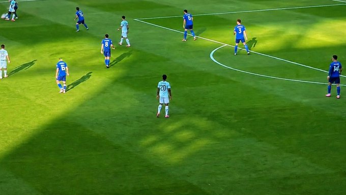 A soccer match is in progress on a sunlit field, with players in light blue and dark blue jerseys—visible numbers include 8, 15, 16, 20, 3, 6, and 14—spread across the pitch near the penalty area arc. A player in a light blue jersey with the number 20 stands prominently in the foreground on the green grass.