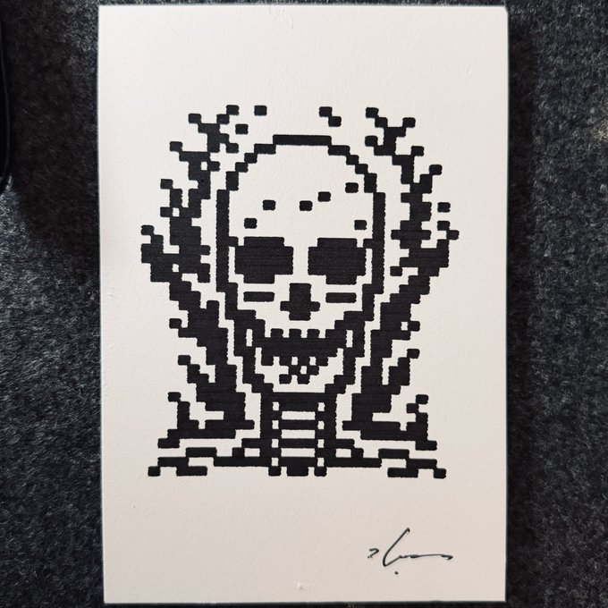 A black-and-white pixel art print depicting a stylized skull, signed in the bottom right corner and resting on a textured dark surface.