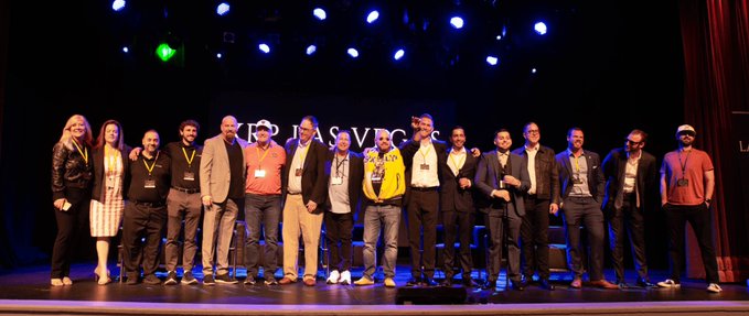 A group photo of approximately 15 people standing on a stage under purple and white lighting, with 'LEADER PASS' visible on the backdrop behind them.