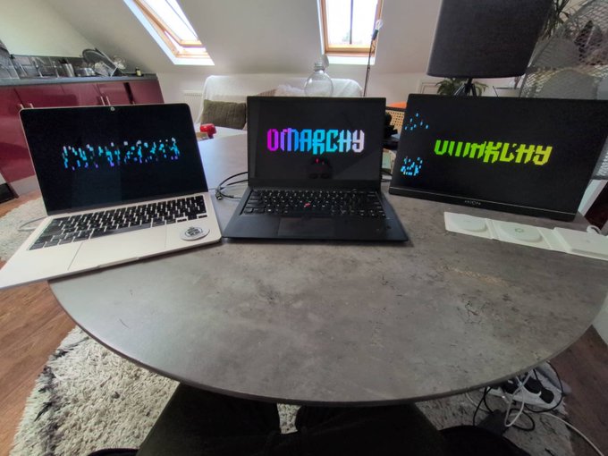 Three laptops sit on a round stone-textured table, each displaying the colorful ASCII art word "OMARCHY" on its screen.