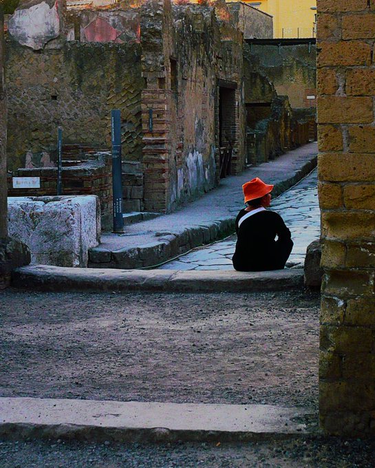 A moody photograph of a person wearing a bright orange hat sitting on the cobblestones of a narrow, weathered street, possibly in an ancient ruin or old town.