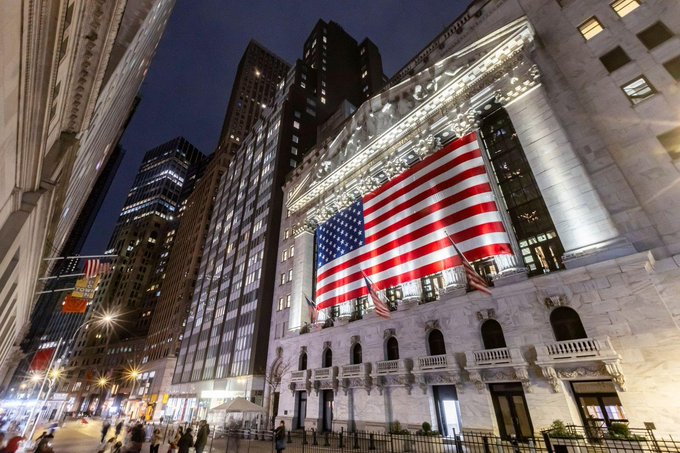 A nighttime view of the New York Stock Exchange building featuring a large American flag draped across its classical facade, illuminated against the dark sky.