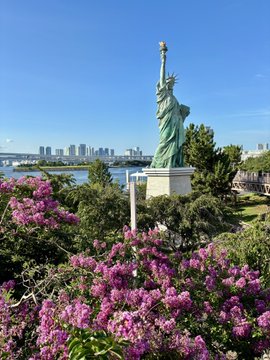 A scenic view of the Statue of Liberty replica (appears to be Odaiba) with vibrant pink crepe myrtle flowers in the foreground and a city skyline across the water.