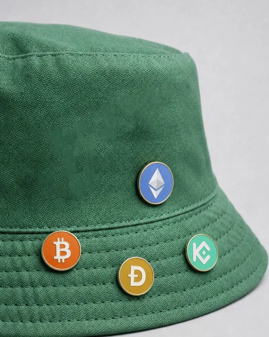 A green bucket hat is adorned with four cryptocurrency-themed enamel pins featuring the Bitcoin (₿), Dogecoin (Ð), Ethereum (Ξ), and KuCoin (K) logos. The caption playfully nods to the post's wordplay: the hat provides literal shade, and the crypto logos symbolize "portfolio protection" through diversified digital asset holdings.