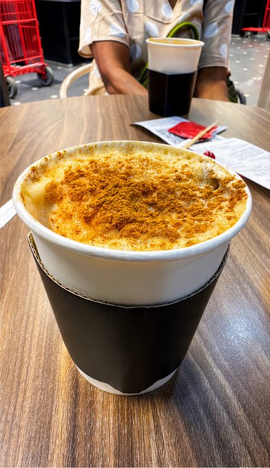 A close-up photograph captures a coffee beverage topped with a thick layer of cinnamon or cocoa powder, served in a paper cup with a black cardboard sleeve on a wooden table, with another drink and a person's lap visible in the background.