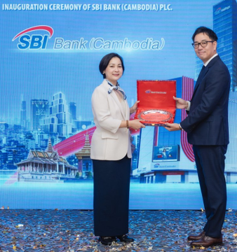 The image shows a man and woman standing next to each other, with the woman holding an open red box while the man holds a small plaque. They are both dressed professionally, wearing suits and ties respectively. The background features a blue wall adorned with a picture of a building, which is part of a promotional event for SBI Bank (Cambodia) in Cambodia.