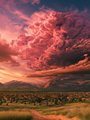 A dramatic landscape photo featuring towering pink and orange-hued cumulus clouds filling the sky over distant green mountains.