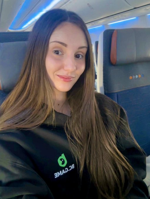 A smiling woman with long brown hair takes a selfie on an airplane, wearing a black sweatshirt featuring the "BC.GAME" logo. The cabin setting with its signature blue overhead lighting and seat-back design confirms she's mid-flight, perfectly matching her playful post teasing her travel destination.