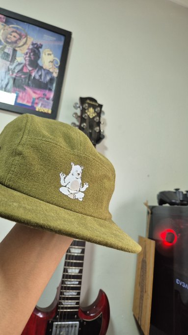 A hand holds up an olive green corduroy cap featuring a small embroidered white bear logo, with a guitar and framed artwork visible in the background.