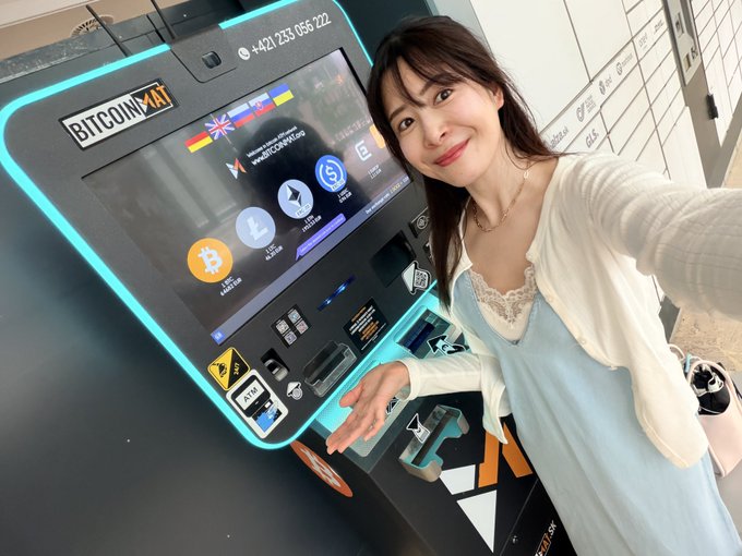 A woman in a light blue dress and white cardigan taking a selfie while standing next to a 'BITCOIN ATM' machine displaying various cryptocurrency logos including Bitcoin and Litecoin.