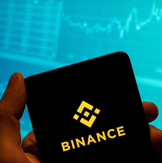 A close-up of a hand holding a smartphone displaying the yellow Binance logo, with a blurred background showing financial charts on a blue screen.