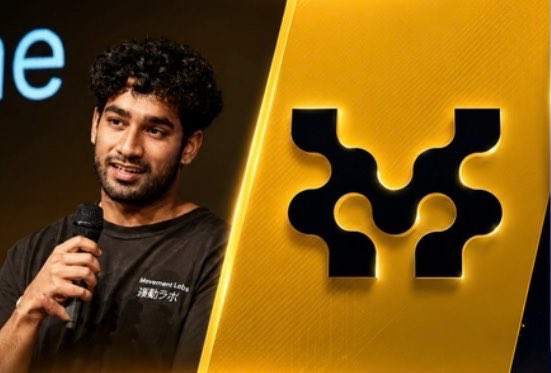 A man with curly hair speaks into a microphone next to a yellow display featuring a black four-pointed logo, with his shirt showing 'Innovation Labs' in English and Japanese.