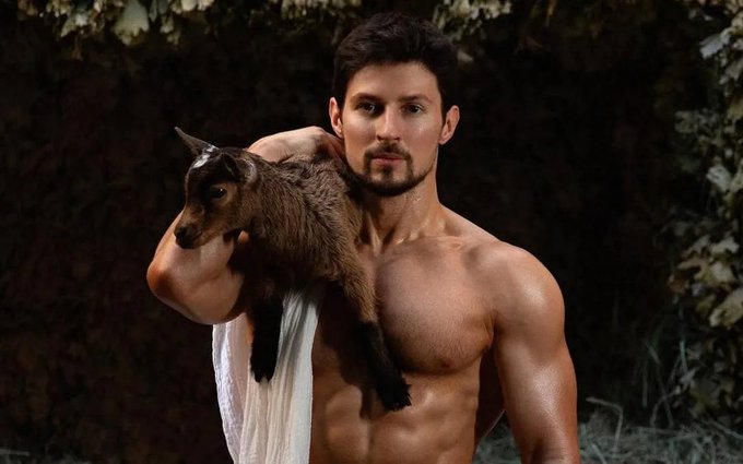 A muscular, bearded man with no shirt holds a small brown goat over his shoulder in a forest setting, dramatically embodying the rugged, "looksmaxxing" aesthetic the post references. The image pairs the man's chiseled physique and brooding expression with the quirky addition of a farm animal, creating a humorous yet intense visual.