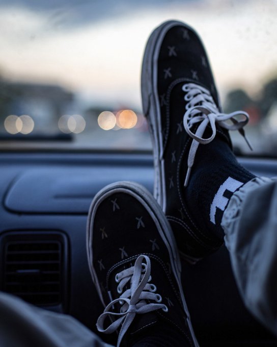 A pair of black Vans sneakers with white laces rests casually on a car dashboard at dusk, with blurred city lights visible through the windshield.
