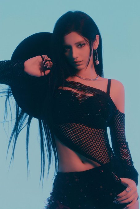 A young woman with long dark hair poses against a light blue background, wearing a black mesh top, dark skirt, and holding a black hat behind her head.