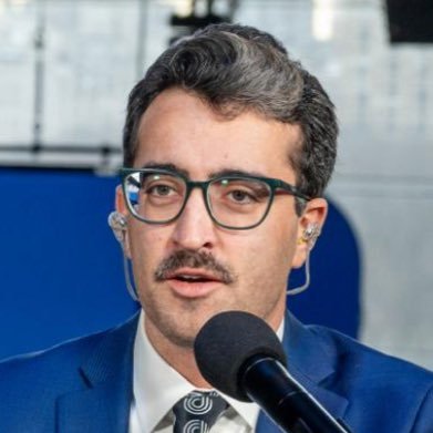 A man with gray hair, glasses, and a mustache speaks into a microphone while wearing a blue suit and tie.