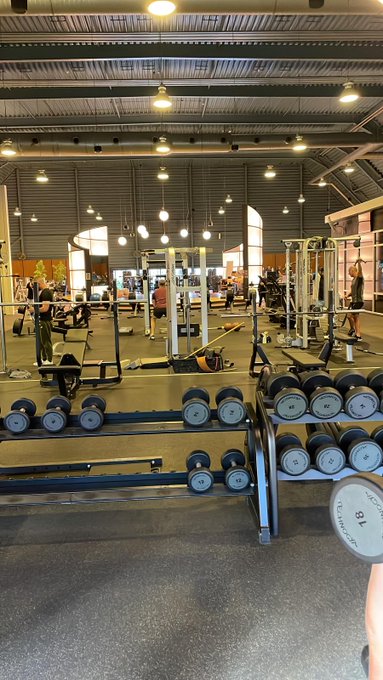 An interior view of a gym showing rows of dumbbells on racks in the foreground, with workout equipment and people exercising in the background under industrial ceiling lights.