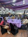 A small, blurry, pixelated image showing a crowded room with people sitting at round tables covered with purple tablecloths, possibly at an event or banquet.