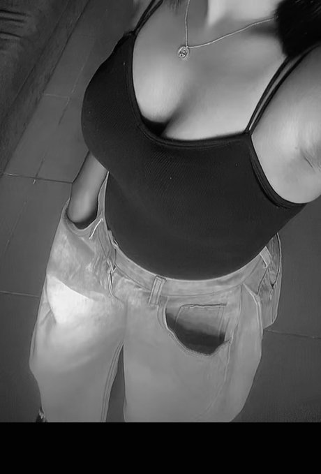 A black and white mirror selfie showing a person wearing a black tank top, light-colored jeans, and a pendant necklace.