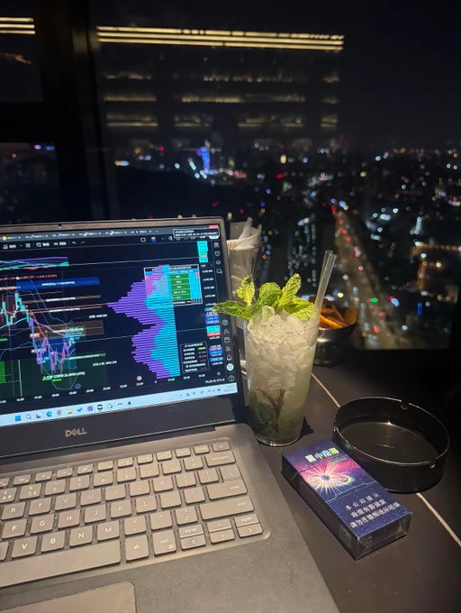 The image features an open laptop computer with a cup of tea, mint leaves, and a cigarette nearby. The scene takes place at night, with a city skyline visible through a window. The focus of the image seems to be on the trading process rather than the financial gains or losses. It emphasizes the importance of patience, proper positioning, and strategic decision-making in trading.