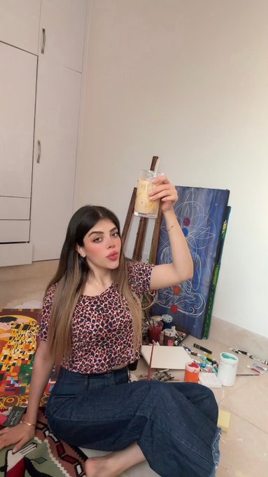 A young woman sits on the floor in what appears to be an art studio, holding up a drink toward the camera while a colorful abstract painting rests on an easel beside her. The post advertises an NFT artwork titled "Noctilume," listed on @objktcom for 30 xtz with an edition of 4/5, tying the artistic setting to the digital art being promoted.