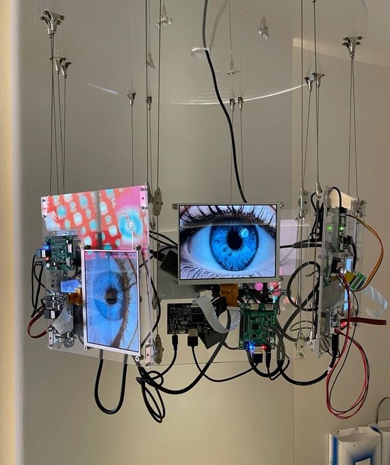 An electronic art installation featuring suspended circuit boards, wires, and screens displaying images of blue eyes and colorful patterns hangs from a ceiling.