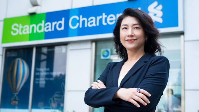 A professional headshot of a woman with dark hair in a black blazer, smiling with arms crossed in front of a Standard Chartered Bank branch with its green and blue signage visible.