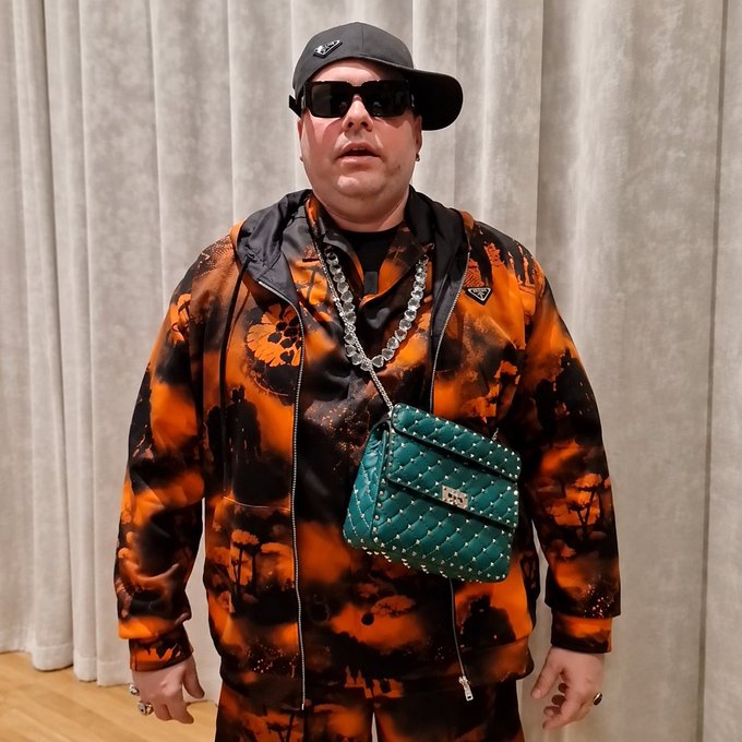 A man in an orange and black tie-dye tracksuit, sunglasses, and a black cap stands in front of gray curtains, accessorized with a large silver chain and a green studded crossbody bag. The image connects to the post by depicting HEX and PulseChain founder Richard Heart, whose recent arrest coincides with a sharp ~44% drop in the $PLS token over the past day.