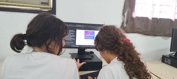 The image shows two people sitting at a desk, looking at a computer screen together. They are both wearing white shirts and appear to be engaged with an online learning platform or tutorial. The background of the image features a window that allows natural light into the room, creating a comfortable atmosphere for studying or working on computers.