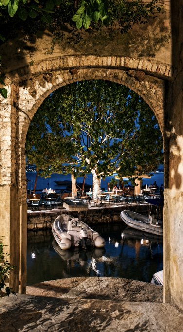 A scenic nighttime view through a stone archway reveals a tranquil harbor with boats, a tree, and distant lights reflecting on calm water.