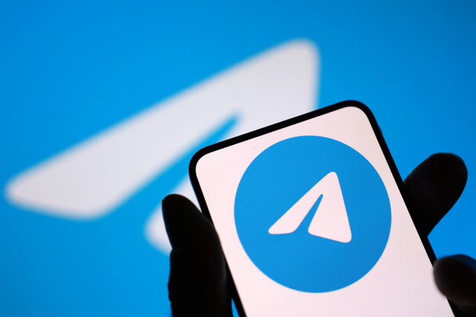 A hand holds up a smartphone displaying the Telegram messaging app logo, set against a larger blurred Telegram logo in the background. The image illustrates the post's skepticism about Telegram's upcoming Gram wallet launch to its massive user base, visually highlighting the platform at the center of the bullish hype.