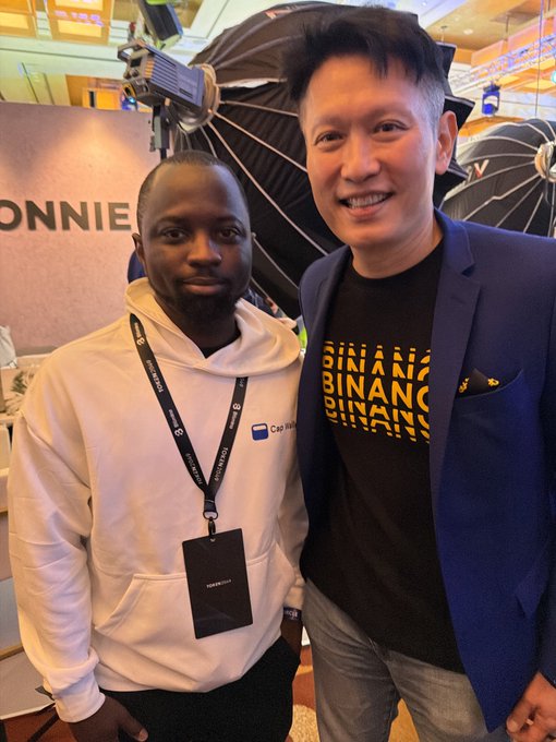 Two men posing for a photo at what appears to be a crypto/blockchain event; one wears a white hoodie with a 'TOKEN2049' lanyard, while the other wears a blue blazer over a 'BINANCE' t-shirt.