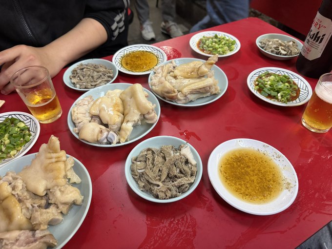 A spread of poached pig's trotters, beef tripe, chopped scallions, and dipping sauces is laid out on a bright red table alongside glasses of Asahi beer and a bottle of Asahi, capturing the hearty, celebratory feast described as "the best of the best Saturday."