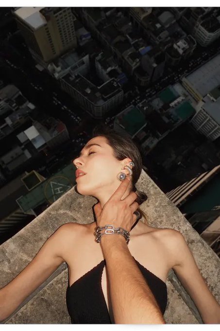A woman with dark hair poses on a high rooftop with her hand on her neck, overlooking a dense urban cityscape below.