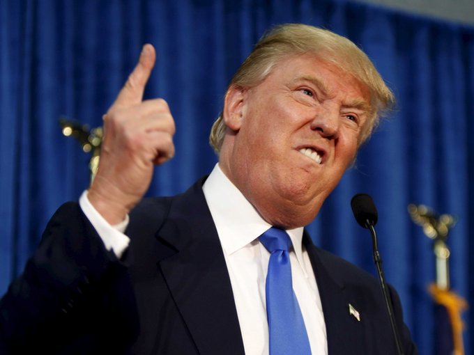 A photograph of Donald Trump in a dark suit and blue tie, smiling and pointing upward with his right index finger while standing at a podium with a microphone.