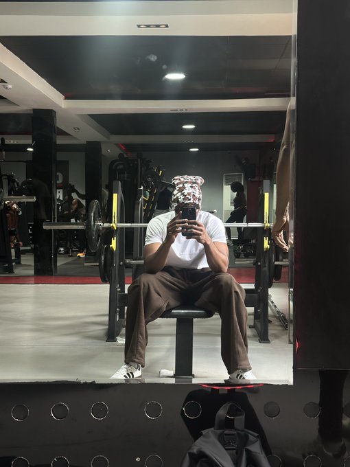 A person sitting on a gym bench taking a mirror selfie while wearing a distinctive animal-print balaclava, a white t-shirt, and brown pants, with a barbell rack in the background.